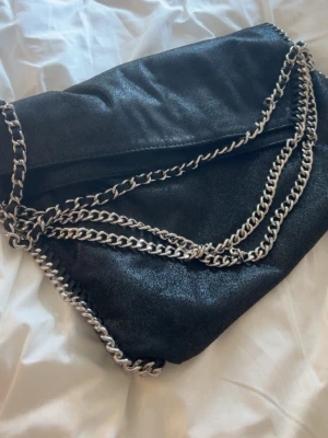 Svart kuvertväska med kedjedetaljer - Snygg svart kuvertväska i mjukt material med silverfärgade kedjor längs kanterna och som axelrem. Väskan har en stilren och modern look med vikbar öppning och passar perfekt till dig som gillar trendiga accessoarer.pris kan diskuteras
