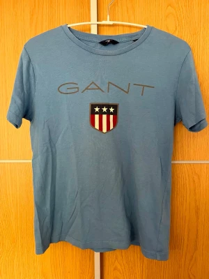 Blå GANT t-shirt med tryck - Snygg blå t-shirt från GANT med tryck framtill och klassisk rund hals. T-shirten har korta ärmar och är gjord i mjuk bomull, perfekt för en avslappnad stil. Stor GANT-logga och sköld med stjärnor och ränder på bröstet.