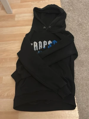 Trapstar hoodie  - Säljer en svart hoodie med huva och dragsko. Framsidan har ett tryck i vitt och blått med texten 'TRAPSTAR'. Hoodien har långa ärmar, ribbade muddar och är gjord i mjukt sweatshirtmaterial. Perfekt för en avslappnad och cool stil.