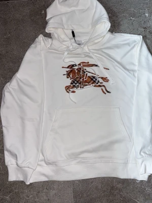 Vit hoodie med rutig hästdesign - Säljer en vit hoodie med stor ficka framtill och huva med dragsko. På bröstet finns ett stort tryck av en häst i brunt och svart rutigt mönster. Hoodien har ribbade muddar vid ärmar och nederkant. Perfekt för dig som gillar unika prints och streetwear.