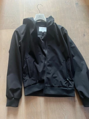 Woolrich jacka oanvänd - Snygg svart oanvänd Woolrichvindjacka med huva och dragkedja framtill. Jackan har två sidofickor med dragkedja och ribbade muddar vid ärmslut och nederkant. Perfekt för blåsiga dagar och enkel att matcha med olika outfits.