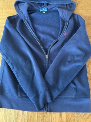 Marinblå hoodie från Polo Ralph Lauren - Snygg marinblå hoodie från Polo Ralph Lauren med dragkedja framtill och huva. Klassisk röd logga broderad på bröstet och två fickor fram. Tillverkad i mjukt material med fleecefoder på insidan för extra komfort.