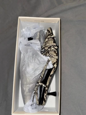 Ormmönstrade pumps med smal klack - Säljer ett par trendiga pumps med ormmönster i beige och svart. Skorna har spetsig tå, smala remmar och en elegant smal klack. Materialet ser ut som konstläder med reptilstruktur. Perfekta för dig som vill sticka ut med din stil.