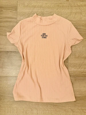 Ribbad t-shirt från Lindex kids 146/152 - Säljer en ribbad ljusrosa t-shirt från Lindex med texten 'AIM FOR THE STARS' broderad framtill. T-shirten har korta ärmar och en lite högre halsringning. Storlek 146/152 kids
