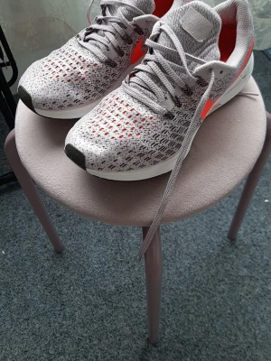 Nike grå/vita sneakers med röd swoosh - Snygga Nike sneakers i grått och vitt med stickad mesh-överdel och röd swoosh på sidan. Skorna har vita sulor och svarta detaljer på tån. Lätta och ventilerande, perfekta för en sportig look. Klassisk låg modell med snörning och bekväm passform.