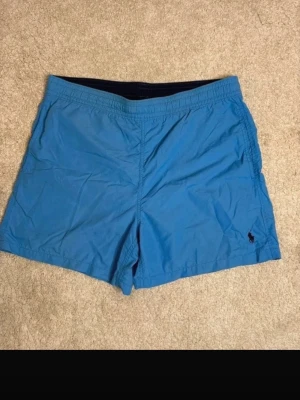 Blå badbyxor från Polo Ralph Lauren - Snygga blå badbyxor från Polo Ralph Lauren med klassisk svart logga broderad på vänster ben. Modellen har elastisk midja och kortare ben, perfekta för stranden eller poolen. Tillverkade i snabbtorkande material för maximal komfort.