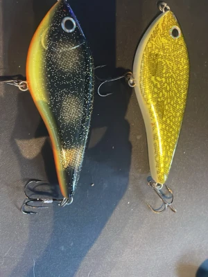 Fiskedrag från Westin, två färger - Två fiskedrag från Westin, ett i svart med glitter och orange/gul kant, det andra i gult med mönster och vit baksida. Båda har trekrokar och är tillverkade i hårdplast. Perfekta för gäddfiske eller annan sportfiske.