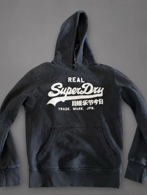 Superdry hoodie - Säljer en svart hoodie från Superdry med stor vit logga och text på bröstet. Hoodien har huva med dragsko och en stor magficka framtill. Perfekt för dig som gillar streetwear och vill ha en skön och snygg tröja med tydlig branding. 7/10 några fläckar, passar xs med