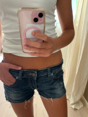 Mörkblå denimshorts med fransar - Säljer ett par mörkblå denimshorts med låg midja och råa fransar längst ner. Klassisk femficksmodell med slitna detaljer och knappgylf. Perfekta för sommardagar och enkel att matcha med olika toppar.