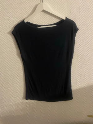 Mörkblå ärmlös topp med bred hals - En stilren mörkblå topp med bred halsringning och ärmlös design. Toppen har en tight passform och är tillverkad i ett mjukt, stretchigt material som känns bekvämt mot huden. Perfekt att matcha med jeans eller kjol för en clean look.