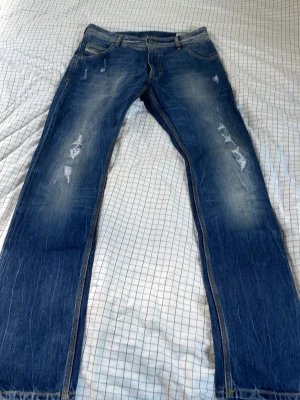 Blå slitna jeans från Diesel - Säljer ett par blå jeans från Diesel med slitningar och snyggt tvättad look. Klassisk femficksmodell med kontrastsömmar och låg midja. Perfekta för dig som gillar en avslappnad och trendig stil.