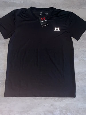 Svart t-shirt från Under Armour - Svart t-shirt från Under Armour med svart logga men blir vit på ljus. T-shirten har en klassisk passform och är tillverkad i ett lätt och funktionellt material som passar perfekt till träning. Enkel och stilren design med rund hals.