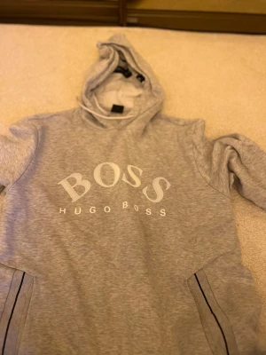 Grå hoodie från Hugo Boss - Snygg grå hoodie från Hugo Boss med stor vit logga på bröstet. Hoodien har huva och två sidofickor med svarta detaljer. Tillverkad i mjukt bomullsmaterial som känns skönt mot huden. Perfekt för en avslappnad och trendig look.