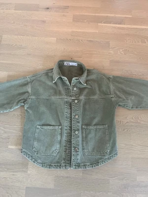 Grön jeansjacka Zara - Olivgrön overshirt i denimliknande material från ZARA. Klassisk krage, tryckknappar framtill och två stora fickor på framsidan. Rak passform och avslappnad stil, perfekt att slänga över en t-shirt.