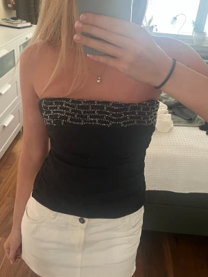 Svart tubtopp med pärldetaljer - Snygg svart tubtopp med glittriga pärldetaljer upptill. Toppen är figurnära och axelbandslös, perfekt för att skapa en trendig look. Materialet känns mjukt och följsamt mot kroppen.