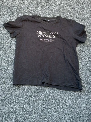 Svart t-shirt med Miami-tryck - Cool svart t-shirt med texten 'Miami Florida NW 98th St' tryckt på bröstet. Klassisk rund hals och korta ärmar. Perfekt för dig som gillar streetstyle och enkel design. [Liten i storleken]