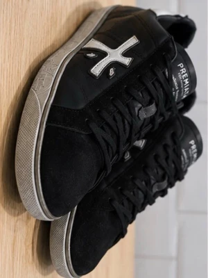 Svarta Premiata sneakers med vit detalj - Snygga svarta sneakers från Premiata med vit logga på sidan och vit häl med texten 'Premiata First Edition'. Skorna har svarta skosnören, vit sula och är tillverkade i en mix av mocka och läder. Perfekta för dig som gillar stilrena och trendiga sneakers.
