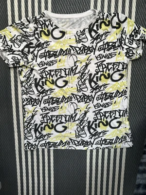 Graffitimönstrad vit t-shirt - Säljer en vit t-shirt med coolt graffitimönster i svart och gult. T-shirten har korta ärmar och en klassisk rund halsringning. Materialet är mjukt och skönt, perfekt för dig som gillar streetstyle och vill sticka ut.
