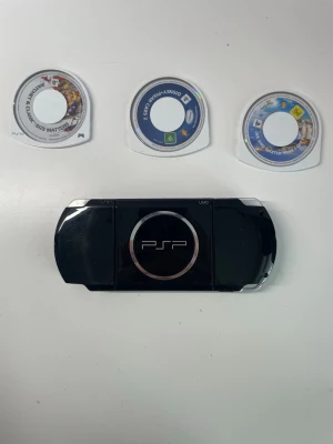 Sony PSP med tre spel - Säljer en svart Sony PSP med tre spel: Ratchet & Clank: Size Matters, Disney Pixar's Cars och Jak and Daxter: The Lost Frontier. Konsolen har en stilren design i svart plast och är perfekt för portabelt spelande. Spelen medföljer i både UMD-format och fodral. Kan erbjuda att bara sälja konsolen för billigare pris