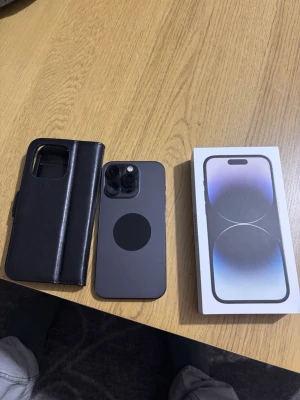 iPhone 14 Pro svart med tillbehör - Säljer en svart iPhone 14 Pro med stilren design och rundade hörn. Telefonen har tre kameror på baksidan och kommer med originalkartong, laddkabel och ett svart fodral i konstläder. Perfekt för dig som vill ha en modern och kraftfull smartphone.