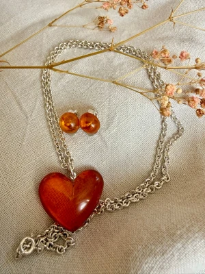 Halsband och örhängen i 925 silver - Vackert set i 925 silver bestående av halsband med hjärtformat hänge samt matchande örhängen. Hänget och örhängena har bärnstensliknande detaljer, men materialet är sannolikt plast/resin och inte äkta bärnsten.  Halsband och örhängen är stämplade 925. Hjärtat har ett mindre ytslitage på ena sidan, vilket inte syns när hänget bärs med den fina sidan fram. Örhängena är något matta, i övrigt fint begagnat skick.  Se bilder för skick och detaljer.  Totalt vikt :20 g