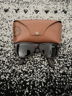 Ray-Ban Wayfarer solglasögon brun - Snygga Ray-Ban Wayfarer solglasögon i mörkbrunt med svarta glas. Klassisk fyrkantig form och detaljer i silver vid fronten. Kommer med ett brunt fodral i läderlook. Perfekta för soliga dagar och ger en cool vibe. Pris kan diskuteras 