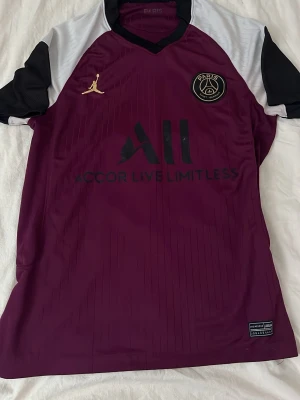 PSG vinröd matchtröja Jordan - Snygg PSG matchtröja i vinrött med svarta och vita detaljer. Tröjan har PSG-logga och Jordan-logga på bröstet samt texten 'Accor Live Limitless' framtill. Tillverkad i ett lätt och ventilerande material, perfekt för fotboll eller träning.