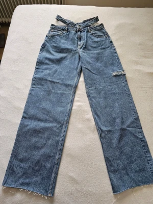 Blå high waist jeans med dubbla linningar - Oanvända ljusblå baggy jeans från Pull&Bear med asymmetrisk criss-cross midja, snygga cut-outs på sidorna, vida ben och rå kant!⚠️VIKTIGT: Står XL på lappen men är extremt små! Passar ca W28/W29 (EU 36/38). 100% bomull helt utan stretch.Mått (platt):• Midja överst: 38 cm• Innerben: 86 cm (Långa/L34)• Midjehöjd: 32 cm till nedre kanten, 35 cm till översta.I absolut nyskick. Rök/djurfritt hem. Fraktar! 📦