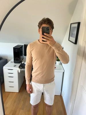 Beige stickad tröja från J. Lindeberg - Snygg beige stickad tröja från J. Lindeberg med rund halsringning och diskret logga på bröstet. Tröjan har långa ärmar och ribbade muddar vid ärmslut och nederkant. Hör gärna av dig vid frågor!