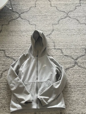 Ljusgrå zip hoodie från Nike - Ljusgrå hoodie med dragkedja från Nike. Klassisk design med huva, två fickor framtill och diskret Nike-logga på bröstet. Tillverkad i mjukt sweatshirtmaterial som är skönt att bära. Perfekt för en avslappnad och sportig stil.