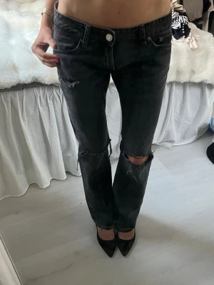 Svarta slitna raka jeans - Säljer ett par svarta raka jeans med slitningar och hål vid knäna. Jeansen har fem fickor, normal midja och en klassisk knappgylf. Perfekta för en avslappnad och trendig look.