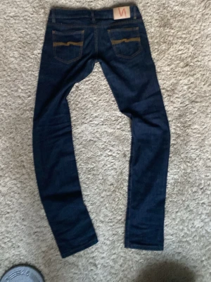 Mörkblå jeans från Nudie Jeans - Säljer ett par mörkblå jeans från Nudie Jeans med klassisk gul söm på bakfickorna och Nudie-logga på läderpatchen. Jeansen har smal passform och är tillverkade i mjukt denimtyg. Perfekta för dig som gillar stilrena och tidlösa jeans.