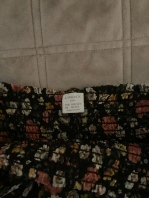 Blommig volangkjol från Lindex - Supersöt kort kjol från Lindex med blommigt mönster i rosa, vit och gul på svart botten. Kjolen har flera volanger och en smockad midja som ger en skön och trendig look. Perfekt för vår och sommar!