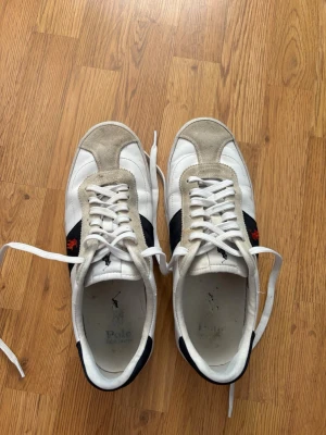 Vita Polo Ralph Lauren sneakers - Snygga vita sneakers från Polo Ralph Lauren med beige mockadetaljer och marinblå sidopaneler. Klassisk låg modell med vit sula och röda logotypbroderier på sidan. Skorna har vita skosnören och är perfekta för en stilren vardagslook. Kan gå ner i pris.