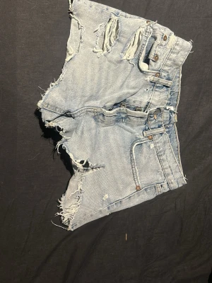 Ljusa denimshorts med slitningar - Säljer ett par ljusa denimshorts med råa och slitna kanter, flera rivna detaljer och klassiska femficksdesign. De är mid waist från Zara. Skriv vid frågor!