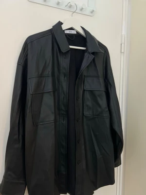 Svart overshirt i skinn från Ellos - Cool svart overshirt från Ellos i skinnimitation med stora bröstfickor och knappar framtill. Overshirten har klassisk krage och en oversized passform som ger en trendig look. Perfekt att slänga över en hoodie eller t-shirt för en edgy stil.