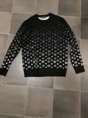 Svart Louis Vuitton tröja med monogram - Snygg svart tröja från Louis Vuitton med klassiskt vitt monogrammönster över hela plagget. Tröjan har rund halsringning och långa ärmar. Perfekt för dig som gillar exklusiv streetstyle och vill sticka ut med en ikonisk design.