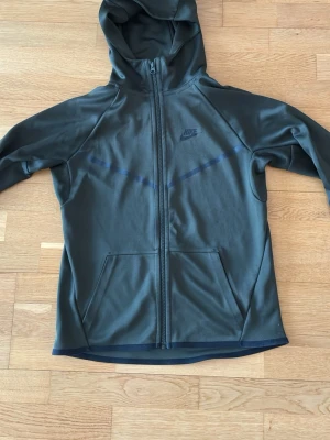 Nike hoodie mörk grön - Snygg mörkgrön hoodie från Nike med hel dragkedja framtill och två fickor. Lite mörkare grön än militärgrön. Tröjan har huva och Nike-logga på bröstet. Hoodien kan användas som ytterplagg i vår med tex en väst. Funkar perfekt till o från gymmet. Rak modell. Sportig design med svarta detaljer. Dragkedjan går högre upp i halsen så hakan kan komma ner under tyger om man drar ända upp. Passar 14-18 år, längd 160-175. Eller S man. Märkt XS men passar mer S Några trådsläpp i armslutet, se bild 4