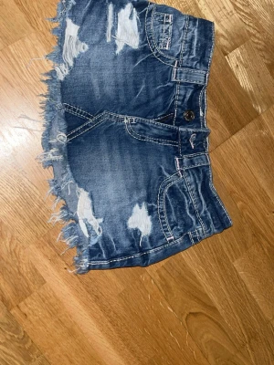Blå jeansshorts med fransig kant - Snygga blå jeansshorts med fransig kant och slitna detaljer framtill. Shortsen har klassiska fickor både fram och bak samt synliga sömmar. Perfekta för sommaren och ger en cool, avslappnad look.