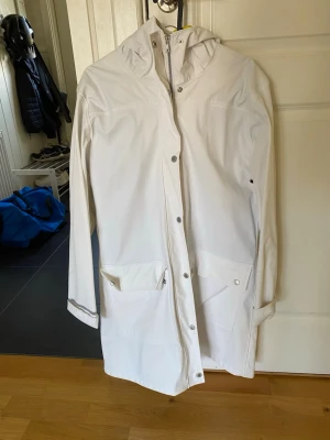 Vit regnjacka med huva - Snygg vit regnjacka med huva och tryckknappar framtill. Jackan har två stora fickor med lock och reflexdetaljer på ärmarna. Lång modell som  skyddar bra mot regn. Perfekt för blöta dagar och enkel att matcha med olika outfits. Regnjackan är tunn. 