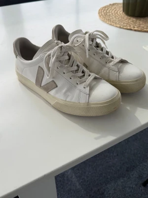 Vita Veja sneakers med beige detaljer - Snygga vita sneakers från Veja med beige V-logga på sidan och matchande beige detaljer. Använda endast i ca en månad, så fortfarande väldigt fräscha. Små tecken på användning finns tex att de är veck i lädret men de är ”vanligt”. Lite missfärgningar på loggan bak ocksåb (se bilder), men i övrigt är de i toppskick.  Lägger till detta pris för att de är rätt så nya, men priset kan diskuteras!   * Storlek: 40 * Färg: Vit * Nypris: 1700 kr   Säljes då de tyvärr inte kommer till användning.