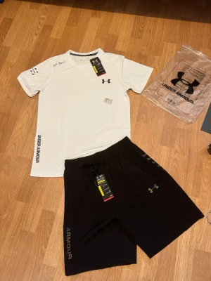 Under armor set storlek m vit\svart - Snyggt träningsset från Under Armour med en vit t-shirt och svarta shorts. T-shirten har tryckt logga på bröstet och text på ärm och nederkant. Shortsen har elastisk midja med snörning och logga på benet. Perfekt för gym eller löpning.