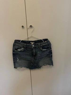 Mörkblå jeanskjol med slitningar - Kort mörkblå jeanskjol med slitna detaljer och rå kant nertill. Klassisk femficksmodell med knapp och dragkedja framtill. Perfekt för en avslappnad och trendig look.