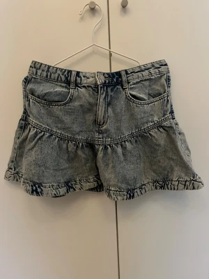 Blå volangkjol i denim - Snygg blå kortkjol i tvättad denim med volangkant och klassiska jeansfickor fram. Kjolen har bälteshällor och en avslappnad loose passform som ger en cool streetstyle-look.