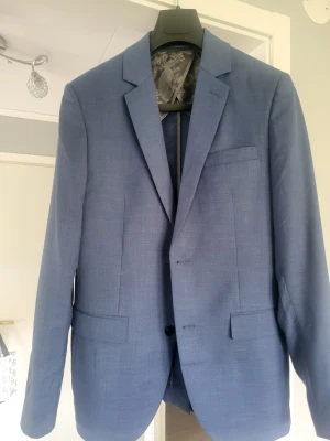 Ljusblå kavaj med klassisk passform - Snygg ljusblå kavaj med två knappar framtill, klassiska slag och tre fickor. Tunn fodring med diskret mönster på insidan. Perfekt för en stilren look. Materialet känns lätt och passar bra till både skjorta och t-shirt ,kostymbyxor ingår ,säljer alltså kostymset.Är ren och fräsch .