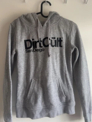 Grå hoodie Dirt Cult San Diego - Snygg grå hoodie från Dirt Cult med svart tryck 'Dirt Cult San Diego' på bröstet. Klassisk känguruficka framtill och justerbar huva med dragsko. Tillverkad i mjukt sweatshirtmaterial, perfekt för en avslappnad streetwear-look.