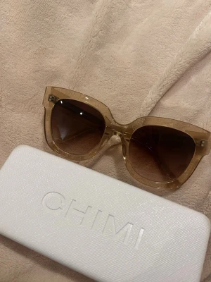  Chimi solglasögon 08 - Säljer ett par stora, fyrkantiga solglasögon från Chimi med transparent beige båge och mörka glas. Kommer i original vit hård väska. Perfekta för dig som vill ha en trendig och stilren look.
