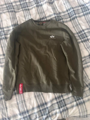 Olivgrön sweatshirt Alpha Industries - Säljer en olivgrön sweatshirt från Alpha Industries med rund halsringning och ribbade muddar. Tröjan har en liten vit logga på bröstet och en röd tag vid nederkanten. Perfekt för en avslappnad streetwear-look.