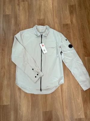 Ljusgrå  overshirt  från C.P. Company - Stilren ljusgrå skjorta från C.P. Company med lång ärm, bröstficka och svart dragkedja framtill. Overshirten har en rundad nederkant och en ikonisk svart detalj på ärmen. Perfekt för dig som gillar modern design.Hör av er vid intresse kan gå ner i pris