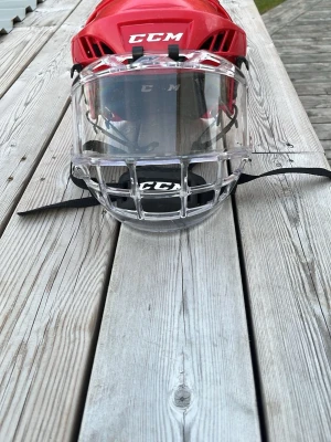 Röd hockeyhjälm från CCM - Röd hockeyhjälm från CCM med transparent visir och galler framtill. Hjälmen har justerbara remmar och är tillverkad i tålig plast med skyddande insida. Perfekt för dig som spelar ishockey och vill ha bra skydd på isen.